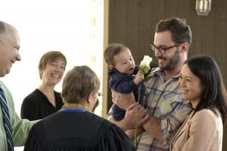 A baby holding a white rose is surrounded by smiling adults at a UU child dedication.