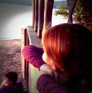 A retro-style photo of a child looking from a porch toward a calm lake while an adult sits below also gazing at lake